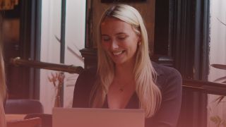 Female recruitment consultant smiling at laptop. There is a pink overlay on the top.